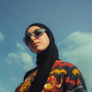 Stylish woman in sunglasses and hijab exuding confidence against a blue sky.